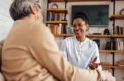 Female companion smiling and conversing with an elderly man in a cozy living room, offering emotional support and friendly conversation.