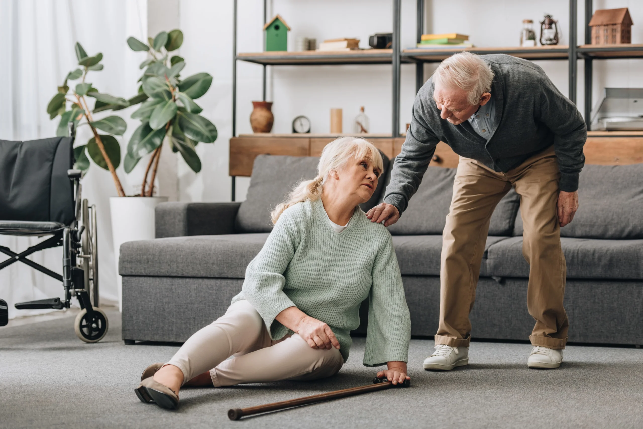 Elderly woman sitting on floor after a fall, supported by senior man, emphasizing fall risk awareness.