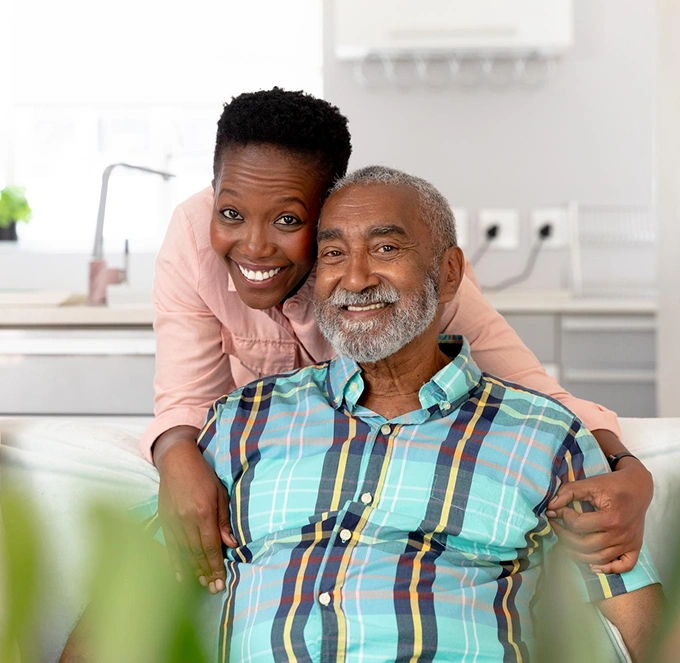 A joyful scene of a woman hugging an elderly man seated on a sofa. She leans over, both smiling warmly.