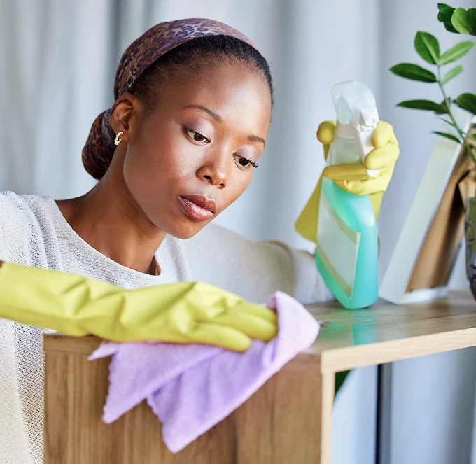 Woman cleaning a wooden shelf with a lavender cloth and spray bottle, wearing yellow gloves. She looks focused, conveying a sense of diligence.