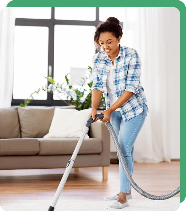 A woman in a plaid shirt and jeans smiles while vacuuming a bright, tidy living room. Sunlight streams through large windows onto a beige sofa.